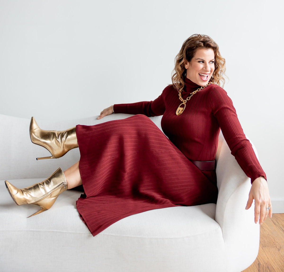 Holly Katz reclining on a white sofa in a burgundy outfit and gold boots